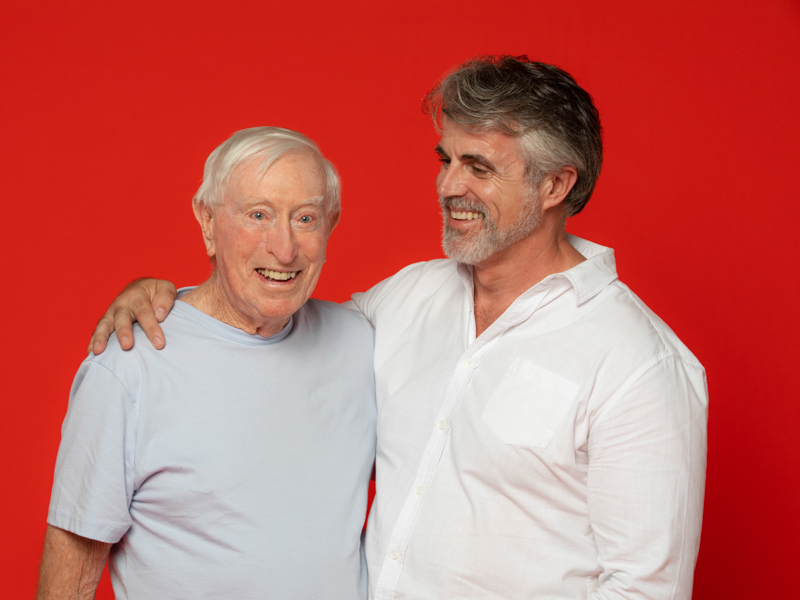 Image of two men looking like father and son with their arms around each other. The son is smiling and looking at this father.